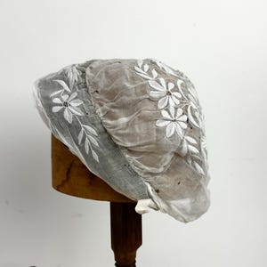 Op de afbeelding: Een doorschijnende, lichtgrijze muts met witte bloemenborduursels. De delicate stof is over een houten vorm gedrapeerd en toont de ingewikkelde details van de geborduurde bloemen en bladeren. De muts is een vintage stijl.
