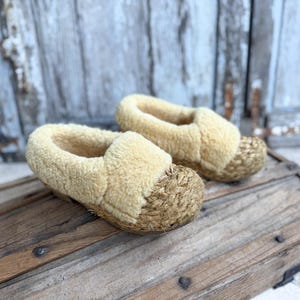 Pode incluir: Um par de chinelos bege com bico tecido e interior macio e fofo. Os chinelos são feitos de um material de cor clara e são projetados para uso interno. Os chinelos estão sobre uma superfície de madeira.