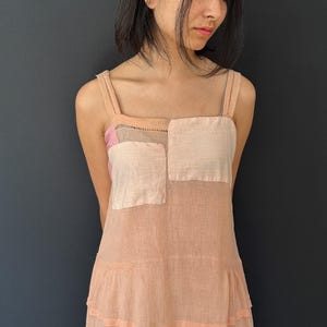 May include: A light peach-colored sundress with a square neckline and thin straps. The bodice features two square patches of light pink and white fabric. The dress has a tiered skirt and is made of a lightweight material.