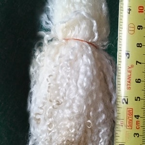 May include: A close-up of a white, fluffy ball of wool yarn, measured against a yellow tape measure. The yarn is approximately 7 inches long.