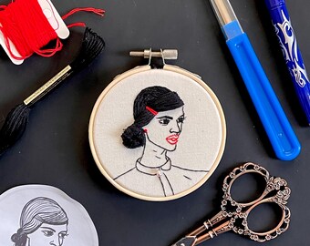May include: A black and white embroidery of a woman's face in a hoop. The woman has black hair, red lips, and a red hair clip. The embroidery is on a white fabric background.