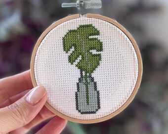 May include: A green monstera leaf in a gray vase, cross-stitched on white fabric and framed in a wooden hoop.