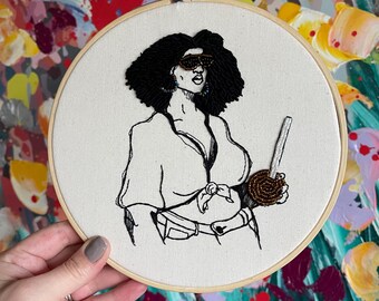 May include: Embroidered hoop art featuring a black woman with an afro wearing sunglasses and a shirt with a tie. She is holding a brown and black beaded object.