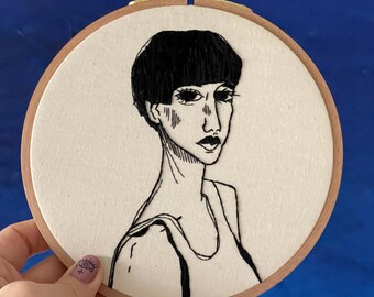 May include: Black and white embroidery of a person with short black hair in a hoop. The person is wearing a sleeveless top.