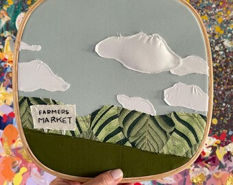 May include: A hand-stitched embroidery hoop with a blue sky, white clouds, green leaves, and a sign that reads "Farmers Market".