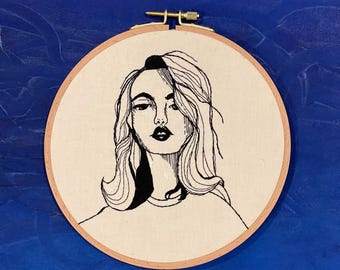 May include: Black and white line drawing embroidery of a woman with long hair in a hoop.