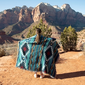 Peut inclure: Une personne debout sur un chemin de terre rouge avec une grande couverture à motifs bleus, bruns et rouges drapé sur ses épaules. La personne est dos à la caméra et regarde une chaîne de montagnes au loin.