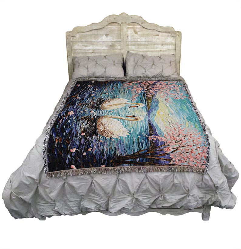 May include: A woven tapestry throw blanket featuring a pair of white swans swimming in a blue lake with pink cherry blossoms in the background. The throw blanket is displayed on a white bed frame with a gray headboard.