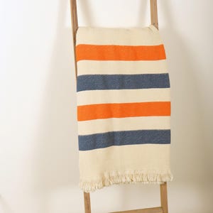 May include: A cream-colored blanket with orange and navy blue stripes is draped over a wooden ladder. The blanket has a fringed edge. The ladder is made of light-colored wood and leans against a white wall.