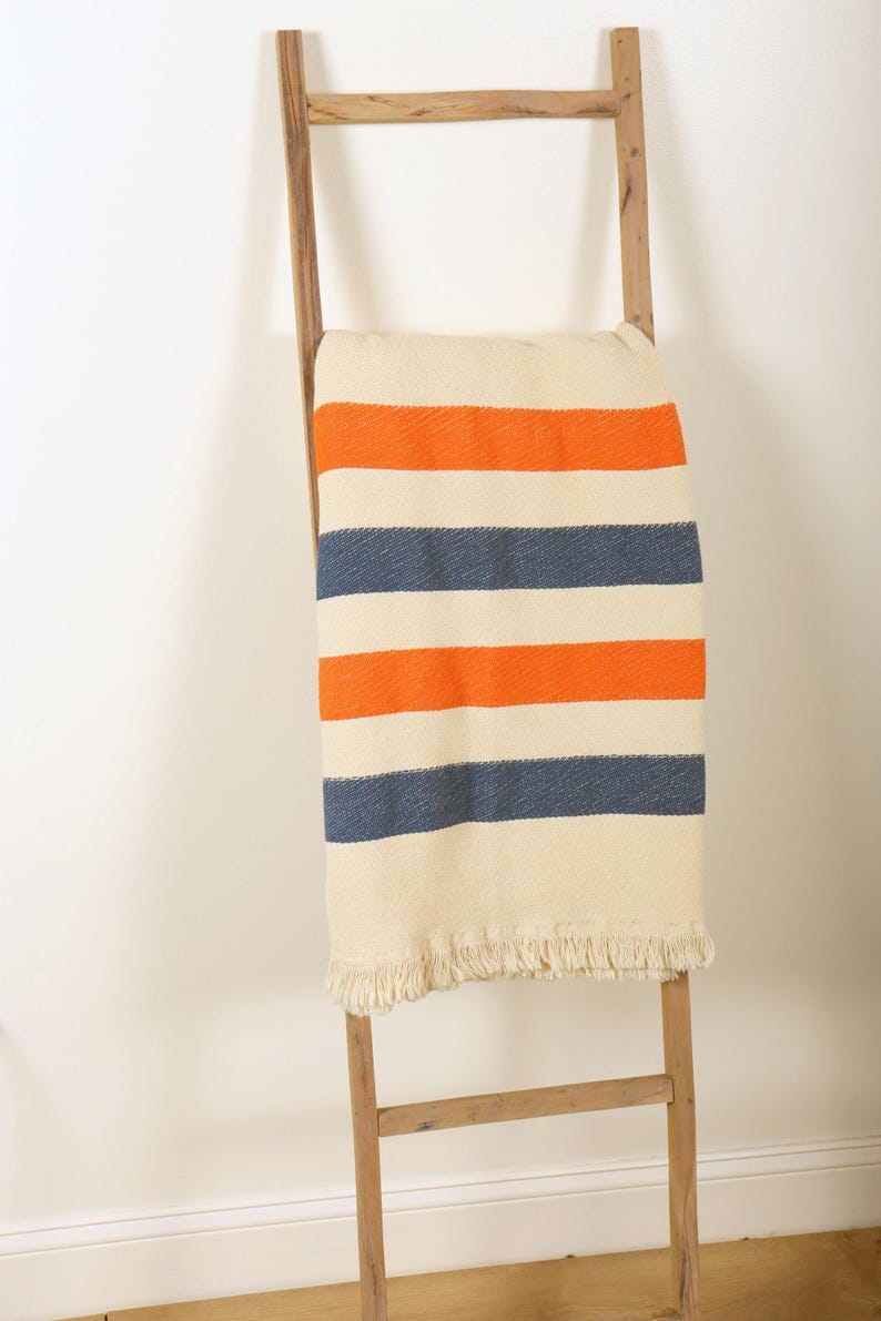 May include: A cream-colored blanket with orange and navy blue stripes is draped over a wooden ladder. The blanket has a fringed edge. The ladder is made of light-colored wood and leans against a white wall.