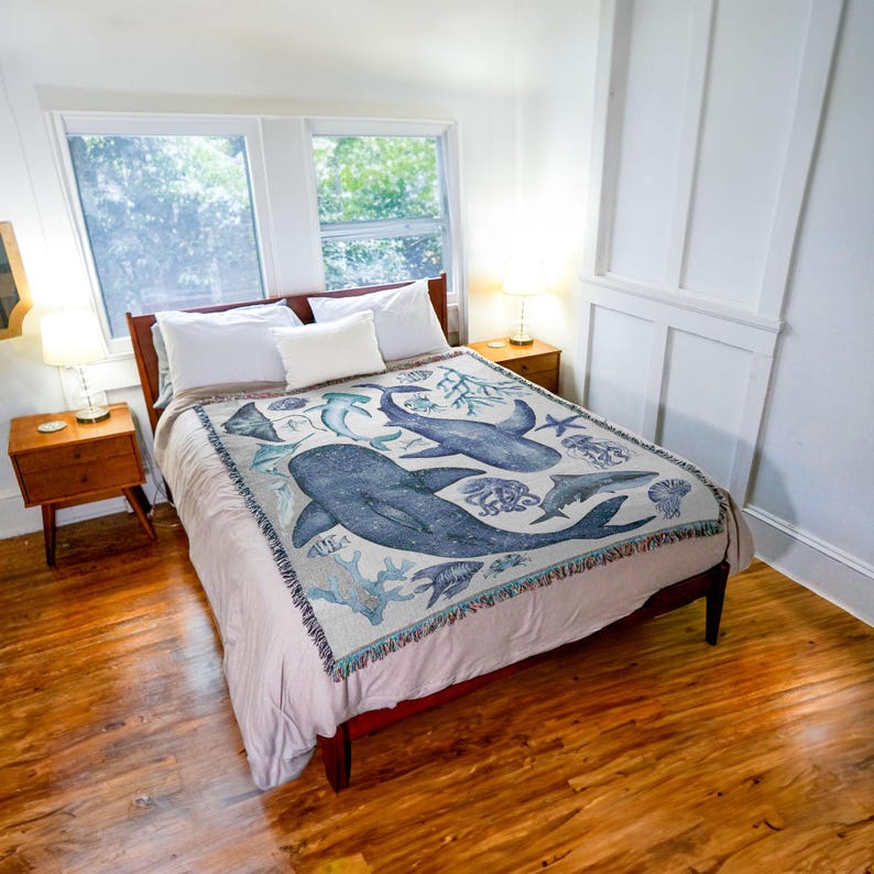 May include: A double bed with a dark wooden frame, white bedding, and a decorative throw. The throw features a blue and white marine life design, including whales, fish, and coral. The room has wooden floors and bedside tables with lamps.
