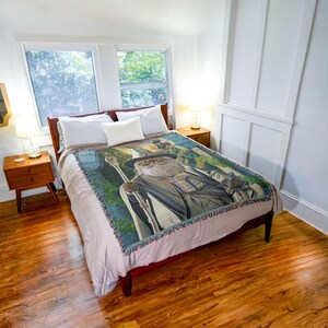 May include: A bed with a brown wooden frame, white pillows, and a decorative throw blanket. The throw features a detailed illustration of a wizard with a staff, in shades of green, blue, and gray. The room has hardwood floors and two bedside tables.
