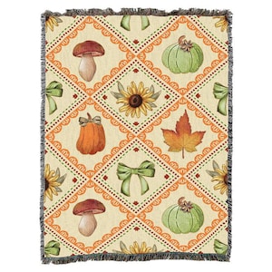 Op de afbeelding: Een geweven plaid met een crèmekleurige achtergrond en een ruitpatroon in oranje. Het ontwerp bevat illustraties van paddenstoelen, groene pompoenen, zonnebloemen, esdoornbladeren en lichtgroene strikken met franjes aan de randen.