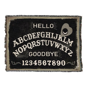 May include: Black and white woven throw blanket with a vintage-style Ouija board design. The board features the letters A-Z, numbers 1-9, and the words "HELLO" and "GOODBYE".