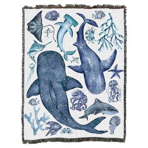 Könnte beinhalten: Eine gewebte Decke mit einem maritimen Thema. Das Design zeigt verschiedene Meeresbewohner wie Haie, Wale und Quallen in Blau- und Türkistönen auf weißem Hintergrund. Die Decke hat einen Fransenrand.