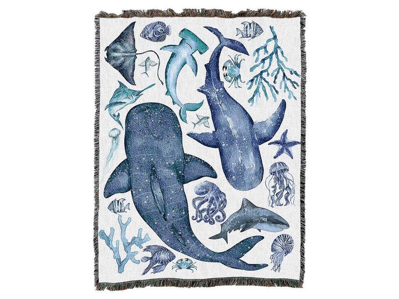 May include: A woven throw blanket with a marine life design. The illustration features sharks, whales, and jellyfish in blue and teal hues on a white backdrop. The blanket has a fringed edge, and is suitable for a sofa or bed.