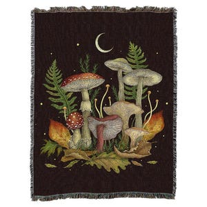 May include: A dark brown woven throw blanket featuring a detailed illustration of various mushrooms, ferns, and autumn leaves. A crescent moon and stars are in the background. The blanket has a fringed edge.