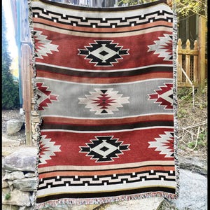 May include: A woven blanket with a Southwestern design. The blanket features a repeating pattern of geometric shapes in red, black, white, and grey. The blanket has a fringed edge and is displayed outdoors.