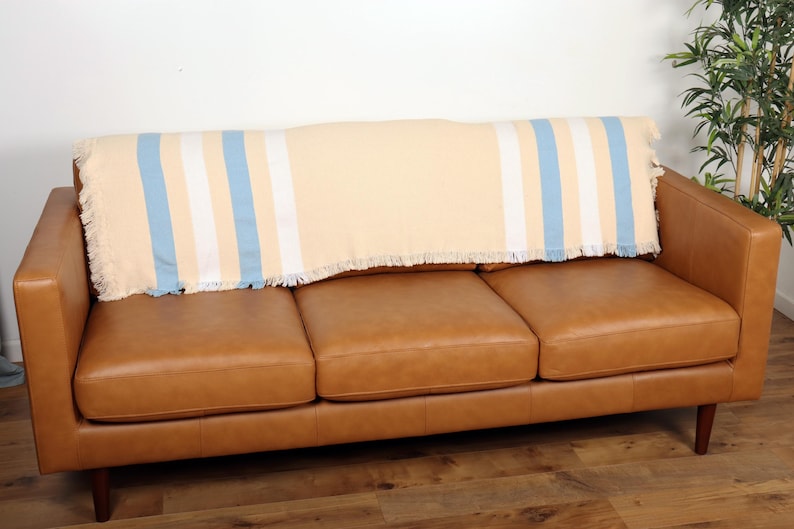 May include: A tan, blue, and white striped throw blanket with fringe is draped over the back of a brown leather sofa. The sofa has three cushions and wooden legs. The background is a plain white wall.