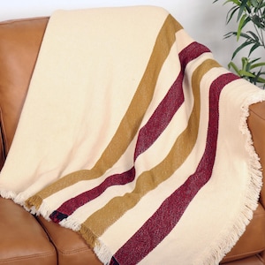 May include: Cream-colored blanket with burgundy and gold stripes, draped over a brown leather sofa. The blanket has a fringed edge. A houseplant is in the background.