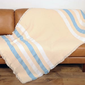 May include: A beige throw blanket with blue and white stripes draped over a brown leather sofa. The blanket has a fringed edge and a soft, inviting appearance. The sofa is set against a wooden floor.