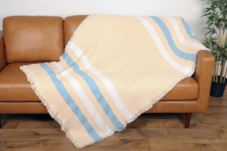 May include: A beige throw blanket with blue and white stripes draped over a brown leather sofa. The blanket has a fringed edge and a soft, inviting appearance. The sofa is set against a wooden floor.