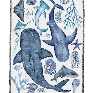 May include: White throw blanket with a marine life design in shades of blue. The design features whales, sharks, jellyfish, and coral. The blanket has a fringed edge.