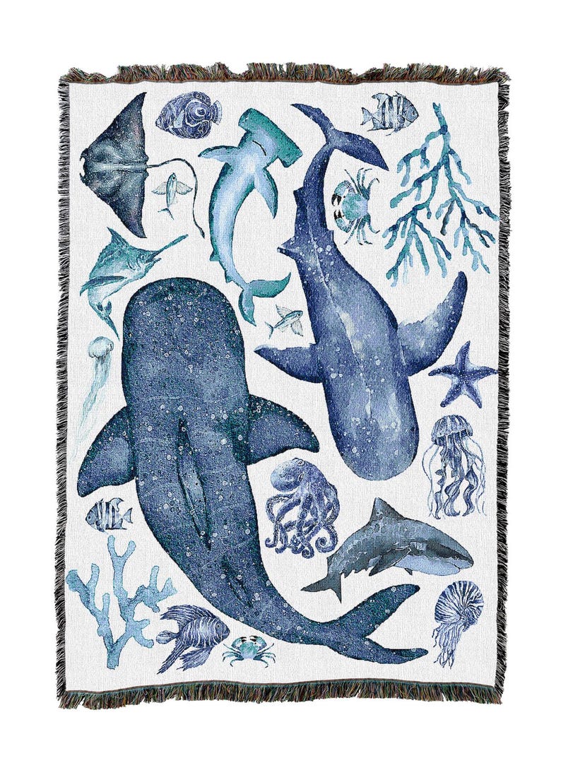 May include: White throw blanket with a marine life design in shades of blue. The design features whales, sharks, jellyfish, and coral. The blanket has a fringed edge.