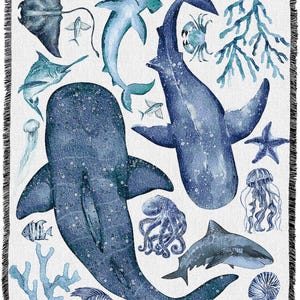 May include: A white throw blanket with a watercolour design of marine life in shades of blue. The design includes sharks, fish, a stingray, and coral. The blanket has a fringed edge.