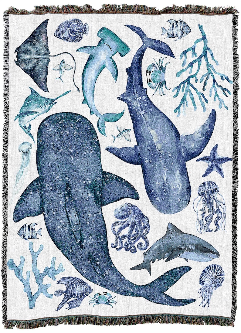 May include: A white throw blanket with a watercolour design of marine life in shades of blue. The design includes sharks, fish, a stingray, and coral. The blanket has a fringed edge.