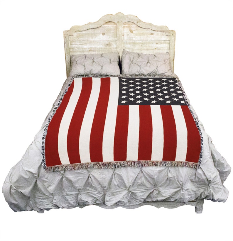 May include: A woven throw blanket featuring the American flag design, with red and white stripes and a black field of white stars. The blanket is draped over a bed with a distressed white headboard and gray bedding.