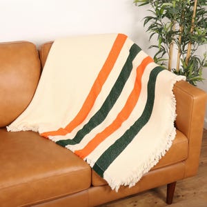 May include: Cream-colored throw blanket with orange and green stripes, fringed edges, draped over a brown leather sofa. The blanket's design features bold, vertical stripes, adding a pop of color to the neutral background.