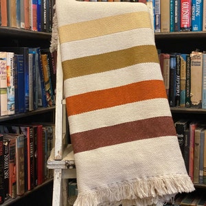 May include: A white throw blanket with brown, orange, and yellow stripes. The blanket has a fringe border and is draped over a white wooden step stool. The text "Bent Creek" is visible above the blanket.