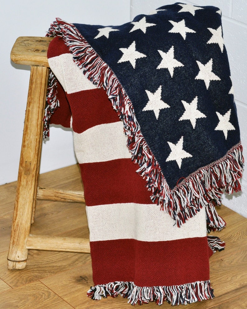 May include: A woven throw blanket featuring the American flag design. The blanket has a navy blue field with white stars and red and white stripes. It is draped over a wooden stool and has fringed edges.