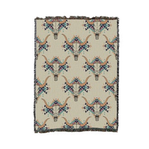May include: A woven throw blanket with a repeating pattern of stylized bull skulls in a geometric design. The pattern is in shades of pink, blue, orange, and brown on a beige background.