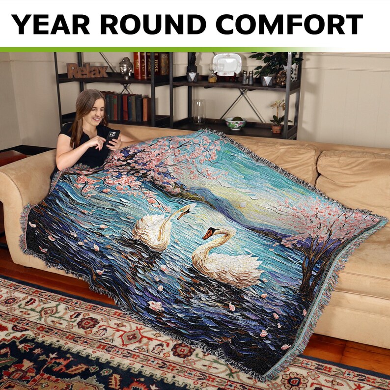 May include: A woven throw blanket with a blue and white design featuring two swans swimming in a lake with pink cherry blossoms on the shore. The throw blanket is draped over a couch.