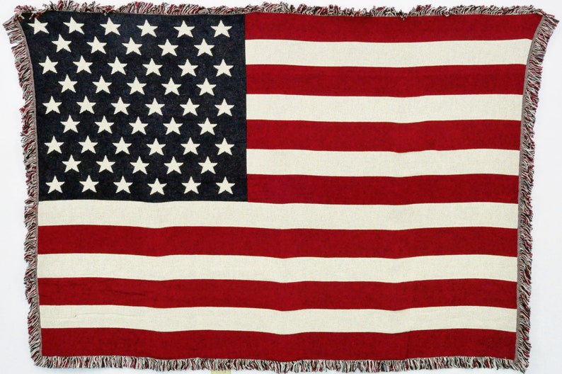 May include: A woven throw blanket featuring the American flag design. The blanket has a navy blue field with white stars in the upper left corner and red and white stripes. The edges are finished with a fringed trim.
