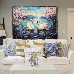 May include: A white throw blanket with a woven tapestry design featuring two white swans swimming in a lake with pink cherry blossoms on the trees. The lake is blue and the sky is a light blue with a sunset in the background.
