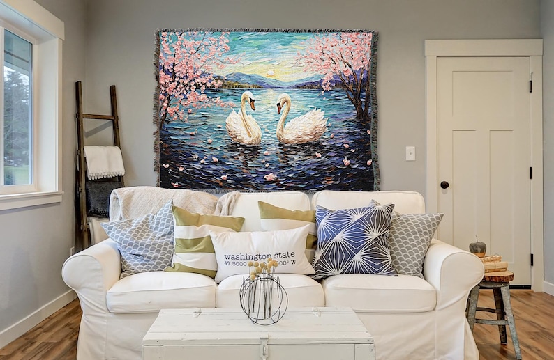 May include: A white throw blanket with a woven tapestry design featuring two white swans swimming in a lake with pink cherry blossoms on the trees. The lake is blue and the sky is a light blue with a sunset in the background.