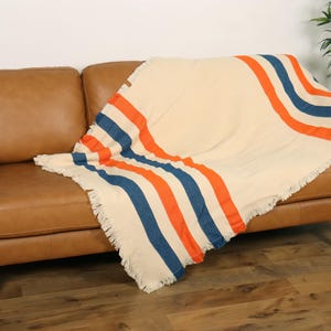 May include: A cream-colored blanket with orange and blue stripes draped over a brown leather sofa. The blanket has a fringed edge. The sofa is in a room with hardwood floors and a plant.