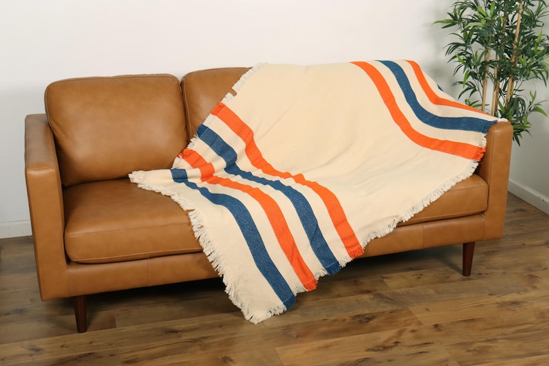 May include: A cream-colored blanket with orange and blue stripes draped over a brown leather sofa. The blanket has a fringed edge. The sofa is in a room with hardwood floors and a plant.