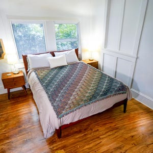 May include: A woven throw blanket featuring a multi-coloured geometric pattern with diagonal stripes in shades of blue, green, orange, and brown, draped over a bed with white pillows and a light-coloured comforter in a bedroom with wooden floors.
