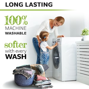 May include: A white washing machine with a woman and a child standing next to it. The text "100% Machine Washable" and "softer with every WASH" is written on the left side of the image. A basket of colorful blankets is in the foreground.