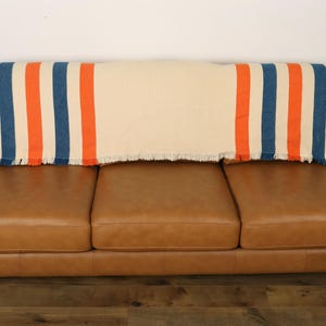 May include: A cream-colored throw blanket with blue and orange stripes is draped over a brown leather sofa. The blanket has a fringed edge. The sofa has three cushions and wooden legs.