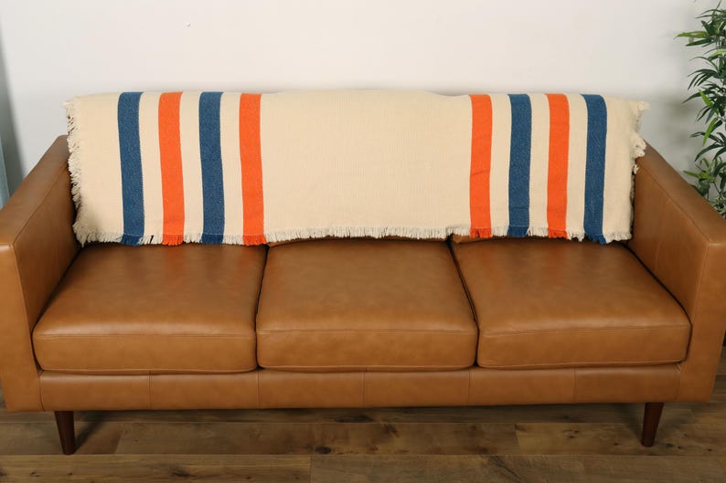 May include: A cream-colored throw blanket with blue and orange stripes is draped over a brown leather sofa. The blanket has a fringed edge. The sofa has three cushions and wooden legs.