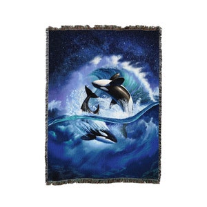 May include: A woven throw blanket featuring a vibrant scene of two orcas leaping through a crashing ocean wave under a starry night sky. The design displays deep blue and white tones with a fringed edge on all sides of the rectangular textile.