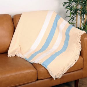 May include: A cream-colored round blanket with blue and white stripes draped over a brown leather couch. The blanket has a fringed edge. The couch is in a well-lit room with a plant in the background.