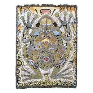 May include: A woven tapestry featuring a large green frog with a bear face in the center. The frog is surrounded by intricate patterns and designs in shades of yellow, green, blue, and red.
