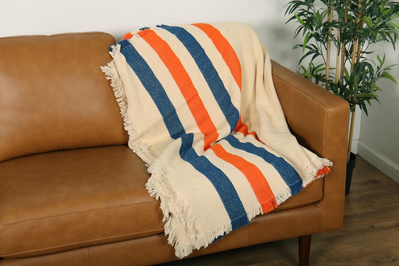 May include: A cream-colored throw blanket with bold orange and blue stripes is draped over a brown leather sofa. The blanket has fringed edges. A houseplant is in the background.