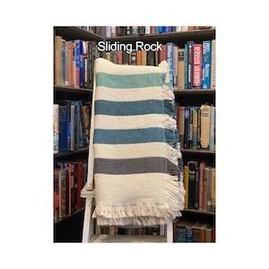 May include: A white, gray, and blue striped throw blanket with fringe on the edges. The blanket is draped over a white wooden chair in front of a bookshelf.